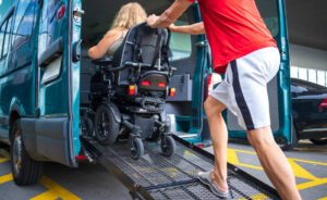 A person in a wheelchair being assisted into a van using a ramp, with someone pushing the wheelchair up the ramp.